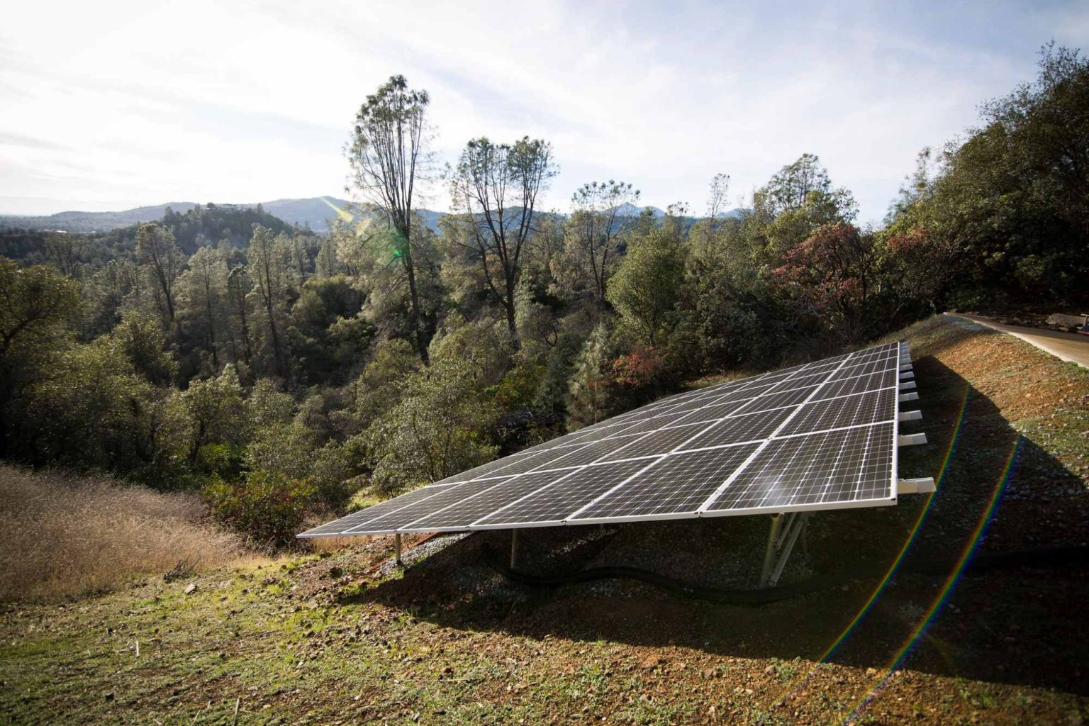 A solar panel array, part of an agricultural solar installation, is set on a grassy hillside surrounded by trees and vegetation, with mountains in the distance under a partly cloudy sky.