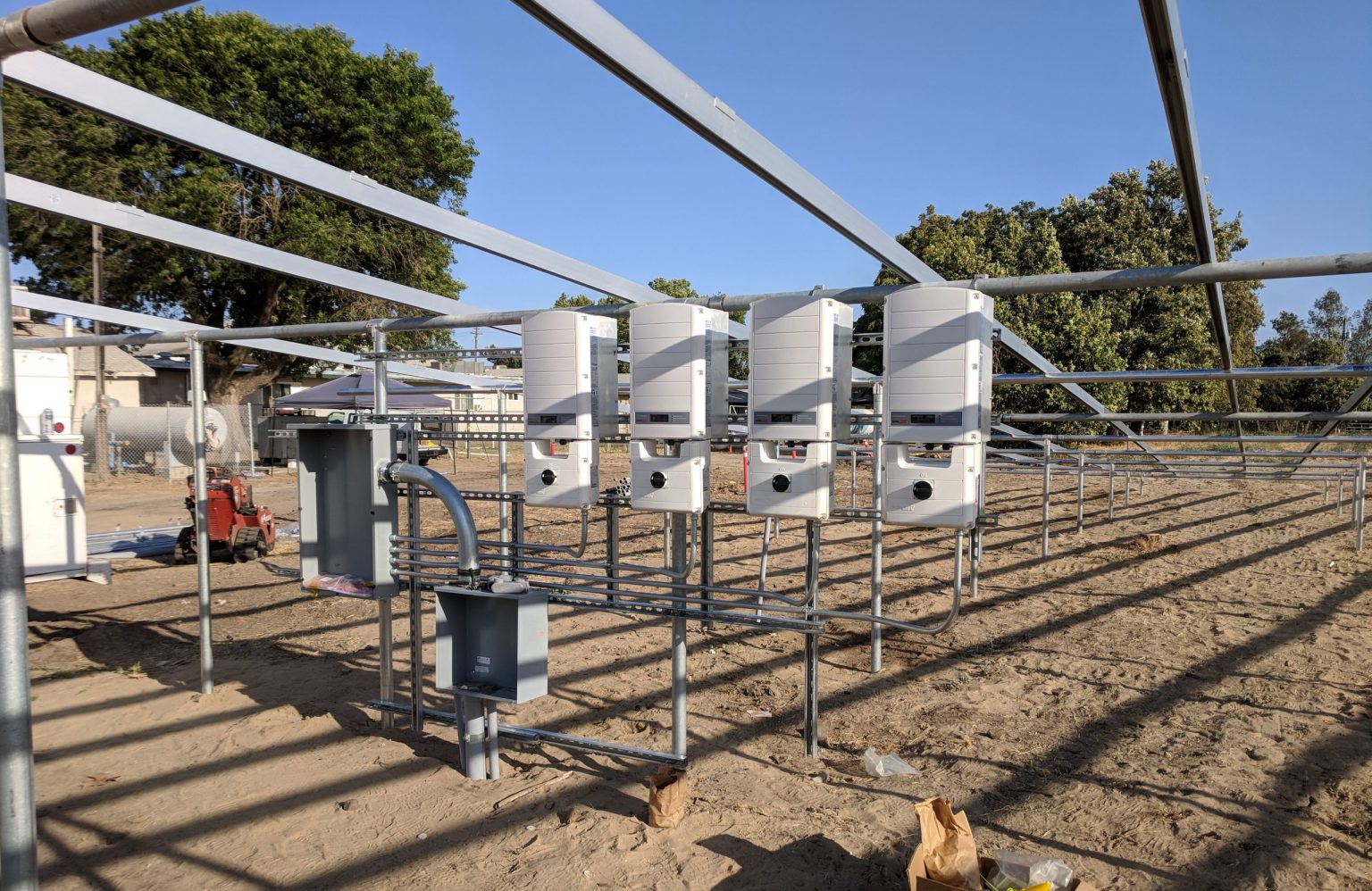 Four electrical inverters are mounted on metal racks in a solar farm under construction, with electrical boxes and conduit visible. This site showcases the importance of Solar System Maintenance. The ground is bare dirt, and trees are in the background.